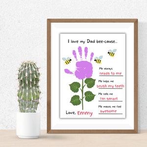 Puede incluir: Una impresi&oacute;n enmarcada con un dise&ntilde;o de flor de huella de mano y el texto "I love my Dad bee-cause..." con razones. La impresi&oacute;n incluye el nombre "Emmy" e ilustraciones de abejas. Un cactus en una maceta blanca est&aacute; a la izquierda.
