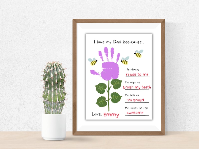 Puede incluir: Una impresi&oacute;n enmarcada con un dise&ntilde;o de flor de huella de mano y el texto "I love my Dad bee-cause..." con razones. La impresi&oacute;n incluye el nombre "Emmy" e ilustraciones de abejas. Un cactus en una maceta blanca est&aacute; a la izquierda.