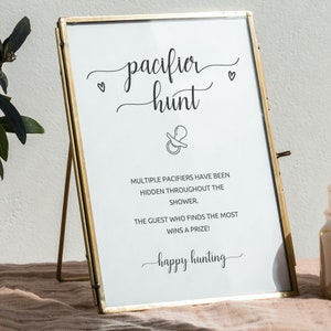 Pode incluir: Uma placa emoldurada com as palavras "pacifier hunt" em escrita elegante, com um gr&aacute;fico de chupeta. A placa inclui o texto "V&aacute;rias chupetas foram escondidas durante o banho. O convidado que encontrar mais ganha um pr&ecirc;mio!"