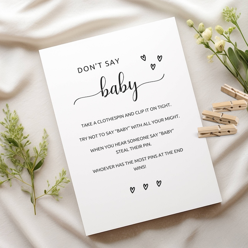 Może przedstawiać: Biała karta z napisem "DON'T SAY baby" w ozdobnej czcionce, z instrukcjami do gry na baby shower. Karta jest ozdobiona małymi ilustracjami serc i jest uzupełniona drewnianymi spinaczami do bielizny.