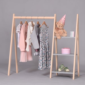 May include: A wooden clothing rack with three shelves. The rack is made of natural wood and has a gray finish. The shelves are also gray. There are several items of clothing hanging on the rack, including a pink dress, a gray sweater, and a floral dress. There is a teddy bear wearing a pink party hat on the top shelf.