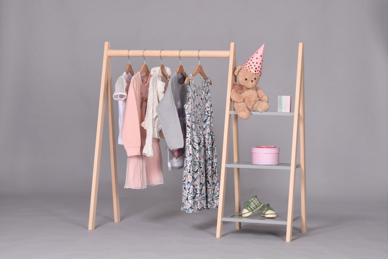 May include: A wooden clothing rack with three shelves. The rack is made of natural wood and has a gray finish. The shelves are also gray. There are several items of clothing hanging on the rack, including a pink dress, a gray sweater, and a floral dress. There is a teddy bear wearing a pink party hat on the top shelf.