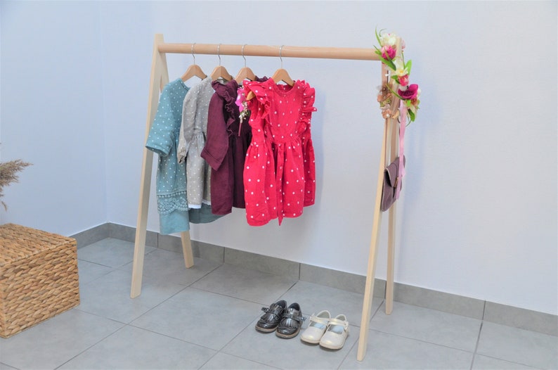 Op de afbeelding: Een houten kledingrek met een natuurlijke afwerking, voorzien van een hangende stang en A-vormige poten. Vier jurken in verschillende kleuren en patronen hangen aan het rek. Twee paar schoenen staan op de vloer voor het rek.