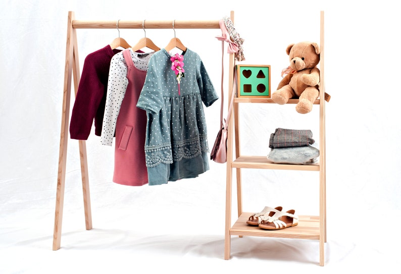 May include: A wooden clothing rack with a hanging rod and two shelves. The rack is holding a burgundy sweater, a white and black polka dot shirt, a pink dress, a blue and white polka dot dress, a pink purse, a teddy bear, a wooden toy with geometric shapes, and a pair of white sandals.