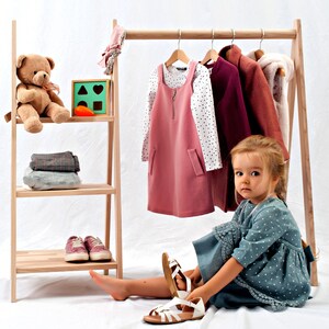 May include: A young child sits on a white floor, putting on a pair of white sandals. The child is wearing a blue and white polka dot dress. Behind the child is a wooden clothing rack with a pink dress, a white long-sleeved shirt with black polka dots, a burgundy jacket, and a brown jacket hanging on it. A wooden shelf with a teddy bear and a wooden toy with geometric shapes is to the left of the rack.