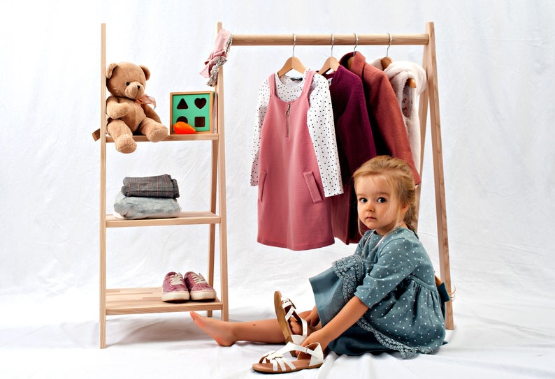 May include: A young child sits on a white floor, putting on a pair of white sandals. The child is wearing a blue and white polka dot dress. Behind the child is a wooden clothing rack with a pink dress, a white long-sleeved shirt with black polka dots, a burgundy jacket, and a brown jacket hanging on it. A wooden shelf with a teddy bear and a wooden toy with geometric shapes is to the left of the rack.