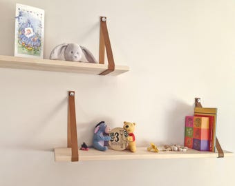 May include: Two wooden shelves with brown leather straps, mounted on a white wall. The top shelf has a card and a stuffed animal, while the bottom shelf has a stuffed animal, a wooden milestone sign that says "3 months", and a stack of books.