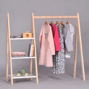 May include: A wooden clothing rack with a shelf on the side. The rack has a white shelf and a wooden frame. The rack is holding a variety of clothing items, including a pink dress, a red dress, a gray sweater, and a white shirt.