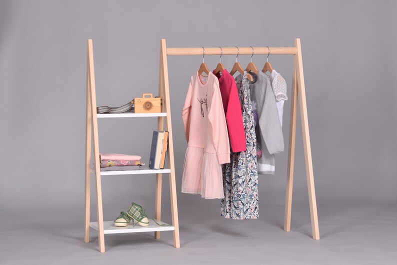 May include: A wooden clothing rack with a shelf on the side. The rack has a white shelf and a wooden frame. The rack is holding a variety of clothing items, including a pink dress, a red dress, a gray sweater, and a white shirt.