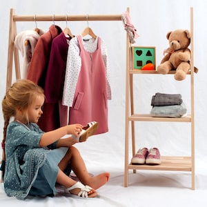 May include: A young girl sits in front of a wooden clothing rack, putting on a pair of white sandals. The rack is filled with clothes, including a pink dress, a white shirt with black polka dots, and a burgundy jacket. A teddy bear sits on a shelf above a stack of folded clothes.
