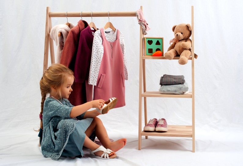 May include: A young girl sits in front of a wooden clothing rack, putting on a pair of white sandals. The rack is filled with clothes, including a pink dress, a white shirt with black polka dots, and a burgundy jacket. A teddy bear sits on a shelf above a stack of folded clothes.