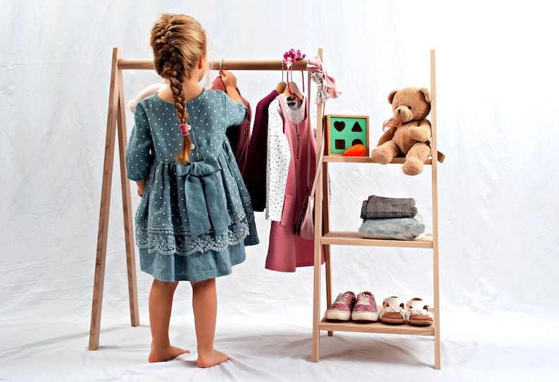 May include: A wooden clothing rack with a child standing in front of it. The rack has a shelf on the bottom and a shelf in the middle. The child is wearing a blue dress with white polka dots. There are clothes hanging on the rack, including a pink dress, a burgundy dress, and a white and black dress.