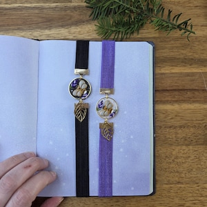 May include: Two bookmarks with purple ribbon and gold accents. One bookmark has a black ribbon and the other has a purple ribbon. Both bookmarks feature a floral design with a white flower and purple accents.