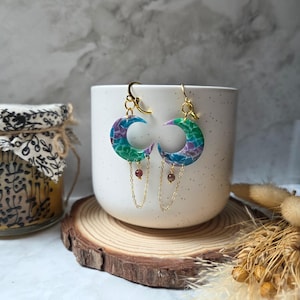 May include: A pair of crescent moon earrings with a colorful, marbled design in shades of blue, green, and purple. The earrings have gold-colored hooks and chains, with small, round, reddish-brown beads dangling from the chains. The earrings are displayed in front of a white mug.