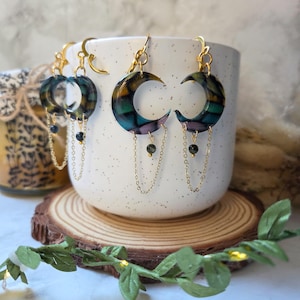 May include: Gold-tone crescent moon earrings with a stained glass effect in shades of green, blue, purple, and yellow. Each earring features a dangling chain with a small black bead. The earrings are displayed in a white ceramic mug.