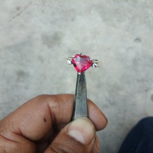 Può includere: Un anello d'argento con una pietra rossa a forma di cuore. L'anello è tenuto da una pinza.