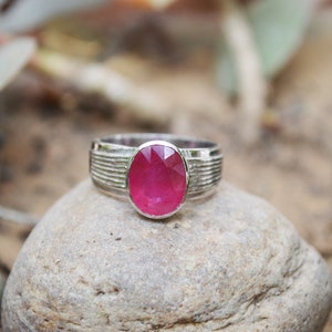 Puede incluir: Un anillo de plata con una piedra preciosa ovalada de rubí rosa brillante engastada en el centro. El anillo tiene una banda texturizada.
