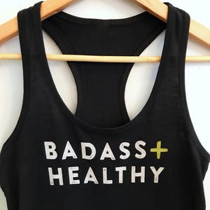K&ouml;nnte beinhalten: Schwarzes Tanktop mit dem Aufdruck "BADASS+HEALTHY" in Wei&szlig; mit einem gelben Pluszeichen.