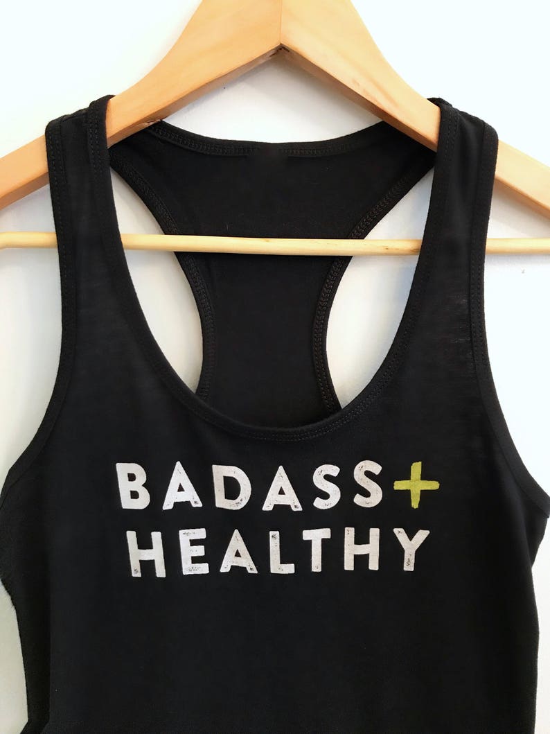 K&ouml;nnte beinhalten: Schwarzes Tanktop mit dem Aufdruck "BADASS+HEALTHY" in Wei&szlig; mit einem gelben Pluszeichen.