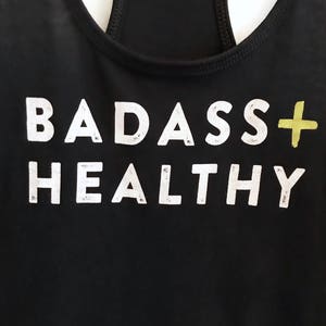 K&ouml;nnte beinhalten: Schwarzes Tanktop mit dem Aufdruck "BADASS+HEALTHY" in Wei&szlig;.