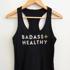 K&ouml;nnte beinhalten: Schwarzes Tanktop mit einem wei&szlig;en Aufdruck, der "BADASS+HEALTHY" mit einem gelben Pluszeichen sagt.