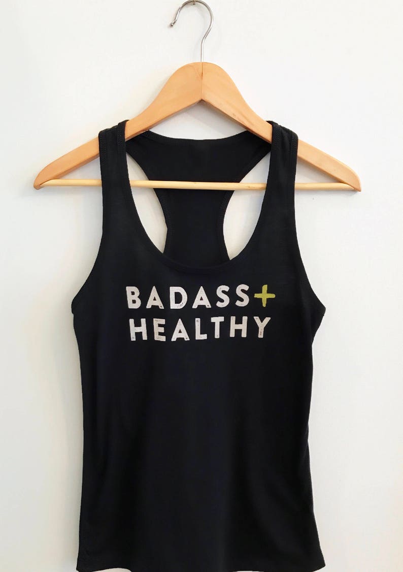 K&ouml;nnte beinhalten: Schwarzes Tanktop mit einem wei&szlig;en Aufdruck, der "BADASS+HEALTHY" mit einem gelben Pluszeichen sagt.