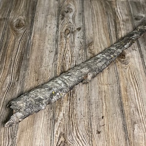 May include: A long, thin piece of driftwood with a rough, gray bark texture. The wood has a natural, weathered look and is lying on a light brown wood surface.