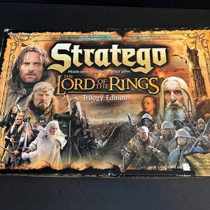 May include: A board game box for Stratego: The Lord of the Rings Trilogy Edition. The box features images of characters from the movie, with the game title in large gold letters. The box is rectangular and has a colorful design.