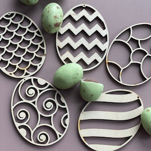 May include: A set of six wooden Easter egg cutouts with different patterns. The cutouts are painted in a light wood colour and have a variety of geometric and abstract designs. The cutouts are arranged on a purple background.