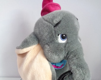 May include: A plush gray elephant toy with a pink hat, blue collar, and large beige ears. The elephant has blue eyes and white accents on its feet. The toy is soft and cuddly, perfect for children.