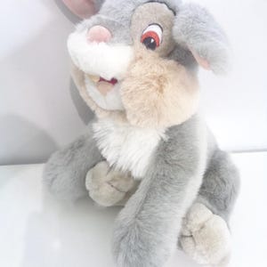 Puede incluir: Un conejo de peluche con pelaje gris, una nariz rosada y ojos grandes y expresivos. El conejo tiene orejas largas con interior rosa y un pecho blanco y esponjoso. El juguete está sentado erguido, con las patas visibles, y tiene una expresión amigable.