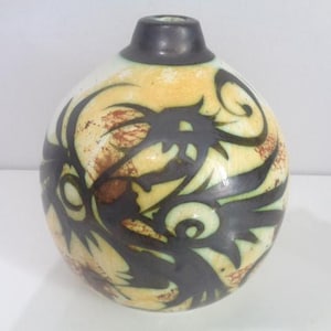 May include: A spherical ceramic vase with a narrow neck and a dark charcoal rim. The body features a cream-coloured base with mottled orange-brown patches and a bold, black, abstract graphic design that wraps around the rounded surface.