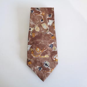 May include: A brown necktie featuring an abstract pattern of scattered, small floral-like shapes in shades of white, yellow, and dark blue. The fabric has a mottled, textured appearance across the entire surface of the pointed tie design.