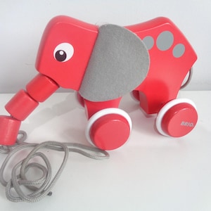 Könnte beinhalten: Ein rotes Holz-Elefanten-Spielzeug zum Ziehen mit einem grauen Filzohr und weißen und roten Rädern. Der Elefant hat einen segmentierten Rüssel und eine graue Schnur. Auf den Rädern steht das Wort "BRIO".
