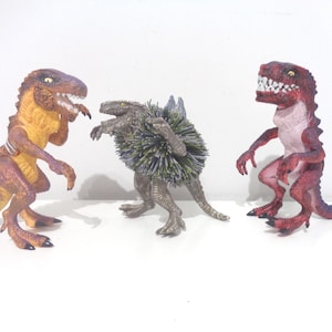 May include: Three toy dinosaurs in a row. The dinosaur on the left is orange and purple. The middle dinosaur is gray with a spiky back. The dinosaur on the right is red and pink.