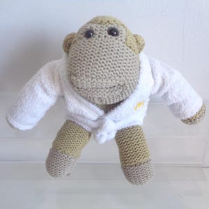 May include: A stuffed monkey toy made of tan knit fabric, featuring dark brown eyes and a small smile. The monkey is wearing a white terry cloth bathrobe with a tied belt at the waist and a small yellow embroidered logo on the left chest area.