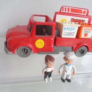 May include: A red toy truck with a smiley face on the side, carrying containers labeled "Fresh Milk" and "Orange Juice." Two small figurines, one in a sailor outfit, stand nearby. The truck has a "Sunny" sign.
