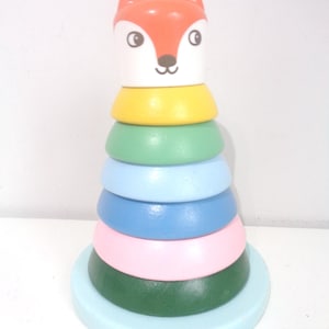 Könnte beinhalten: Ein Holz-Steckspielzeug mit einem Fuchskopf und farbigen Ringen. Der Fuchskopf ist orange und weiß, mit schwarzen Augen und einer kleinen schwarzen Nase. Die Ringe sind gelb, grün, hellblau, dunkelblau und rosa, gestapelt auf einer hellblauen Basis.