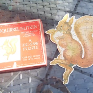 May include: A red box with the words "Squirrel Nutkin" and "A Jig-Saw Puzzle" on it. The box is next to a cut-out jigsaw puzzle of a squirrel with a brown and white tail. The puzzle is 14 inches by 11 inches.