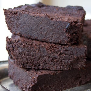 May include: A stack of three dark brown brownies with a slightly textured surface. The brownies are stacked on top of each other, with a silver tray in the background. The brownies appear to be freshly baked.