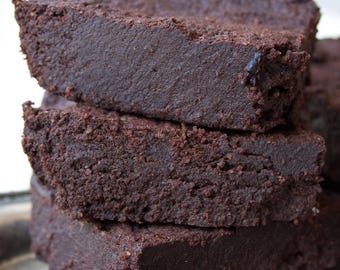 May include: A stack of three dark brown brownies with a slightly textured surface. The brownies are stacked on top of each other, with a silver tray in the background. The brownies appear to be freshly baked.