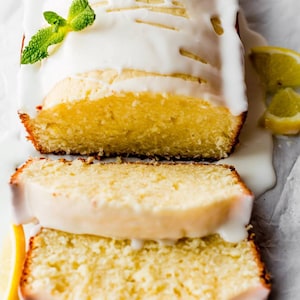 May include: A sliced lemon loaf cake with white icing and a sprig of mint. The cake is light yellow with a golden-brown crust. Lemon slices are placed around the cake on a white surface.
