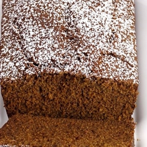 May include: A loaf of freshly baked, brown bread dusted with powdered sugar, with two slices cut and placed in front. The bread is on a white plate, suggesting a homemade or bakery-style treat.