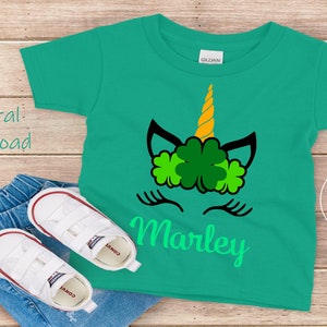 May include: A green t-shirt with a unicorn face design featuring a gold horn and shamrock leaves. The name "Marley" is written in green below the unicorn face.