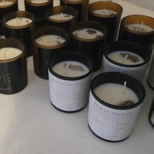 May include: An assortment of black glass candles with white wax and wicks. Some candles have dried botanicals on top. The labels read "Head of The Drum Sweetgrass Braids Natural Soy Candle".