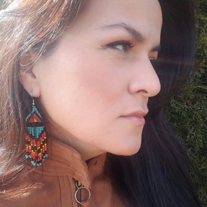 May include: A woman wearing a brown leather jacket with a pair of long, beaded earrings. The earrings are black, red, orange, turquoise, and white.