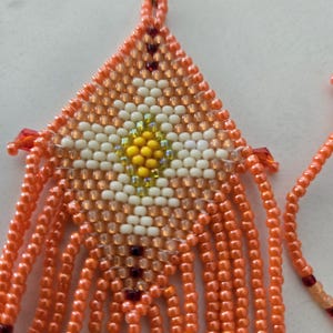 Pode incluir: Close-up de um par de brincos de contas. Os brincos são triangulares com um design floral central em contas brancas e amarelas. As bordas externas e a franja são laranja. Os brincos penduram de um gancho prateado.