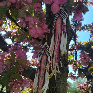 May include: A pair of beaded earrings with a geometric pattern and fringe. The earrings are white, black, pink, and gold. The earrings are hanging from a tree branch with pink blossoms.