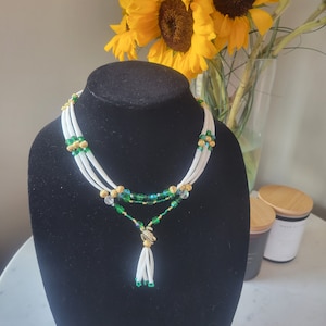 May include: A white and green beaded necklace with a gold turtle pendant and a white tassel. The necklace is layered and features green and clear beads.