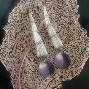 May include: A pair of white and gold shell earrings with purple shell accents. The earrings are hanging from a coral-like background.